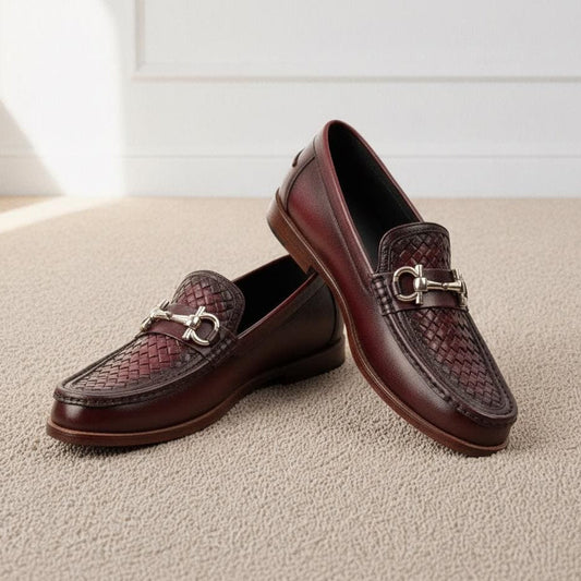 A pair of dark brown leather loafers with a woven pattern on the toe and a silver-tone horsebit detail, displayed on a beige carpeted floor.