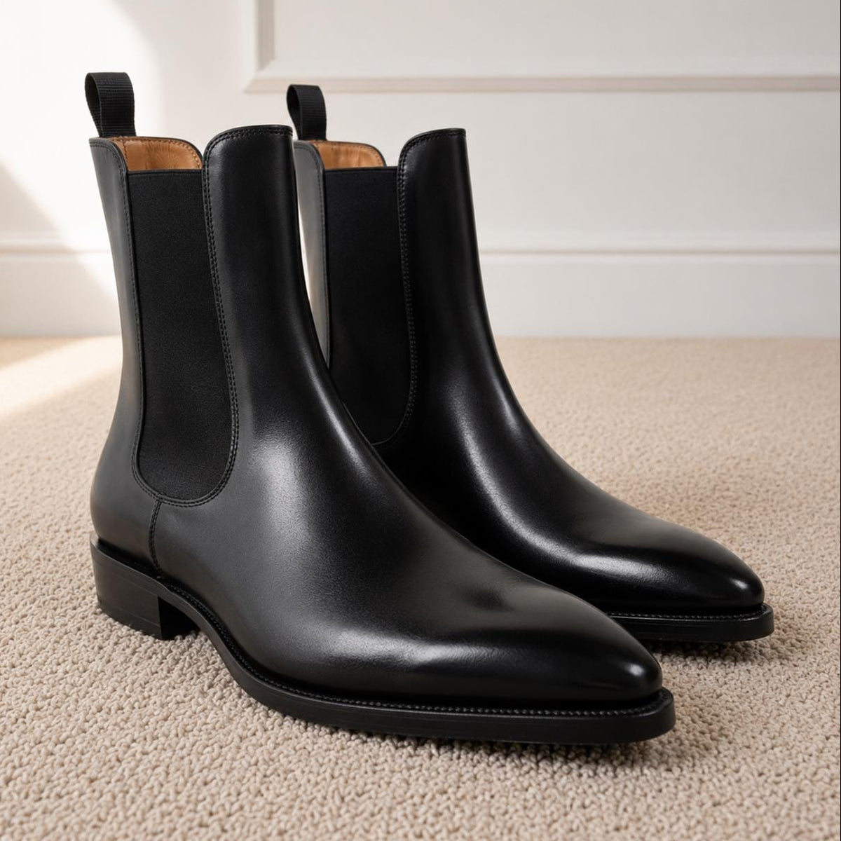 Pair of black polished leather high-shaft Chelsea boots with a pointed toe and black elastic side panels on a neutral carpet